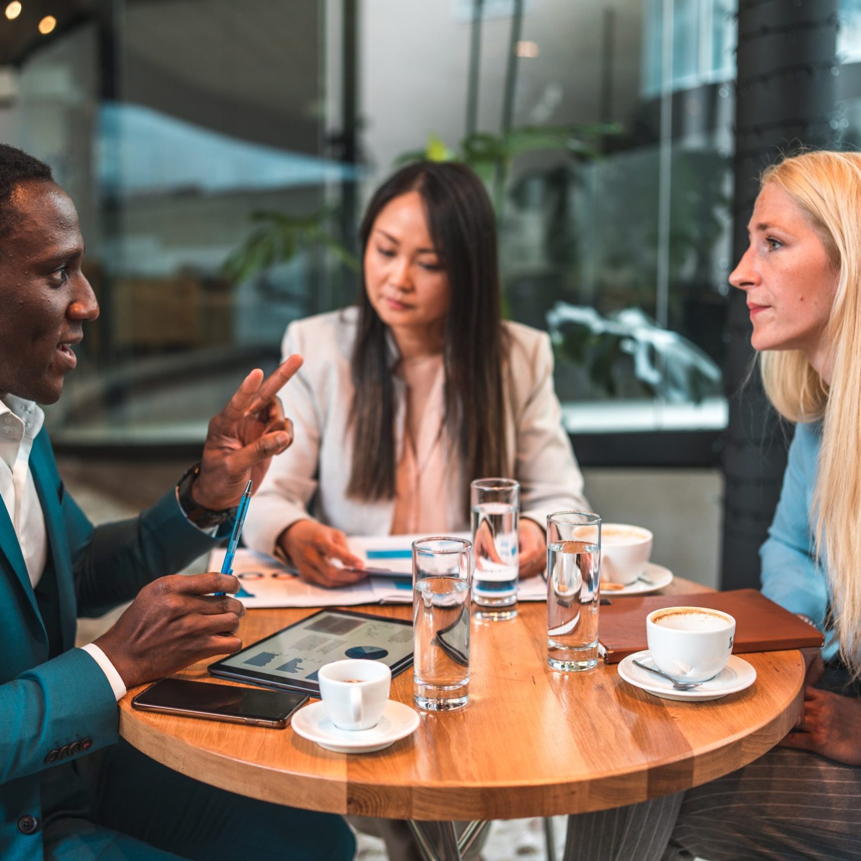 Experience the strength of multi-cultural collaboration as a Black businessman, a Caucasian businesswoman, and an Asian businesswoman gather in a chic cafeteria, engaging in an effective planning session to drive their business forward.